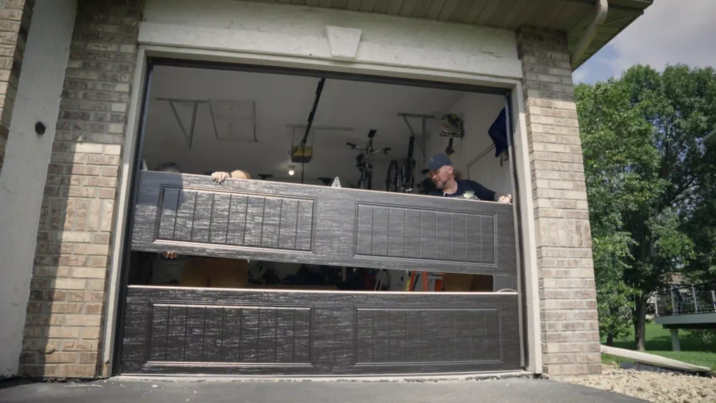 Two installers fit the second section of a new garage door into a brick-framed opening. The bottom panel is already installed, and both panels have a dark brown woodgrain finish with recessed, vertically grooved inserts. Bikes hang inside the garage behind them.